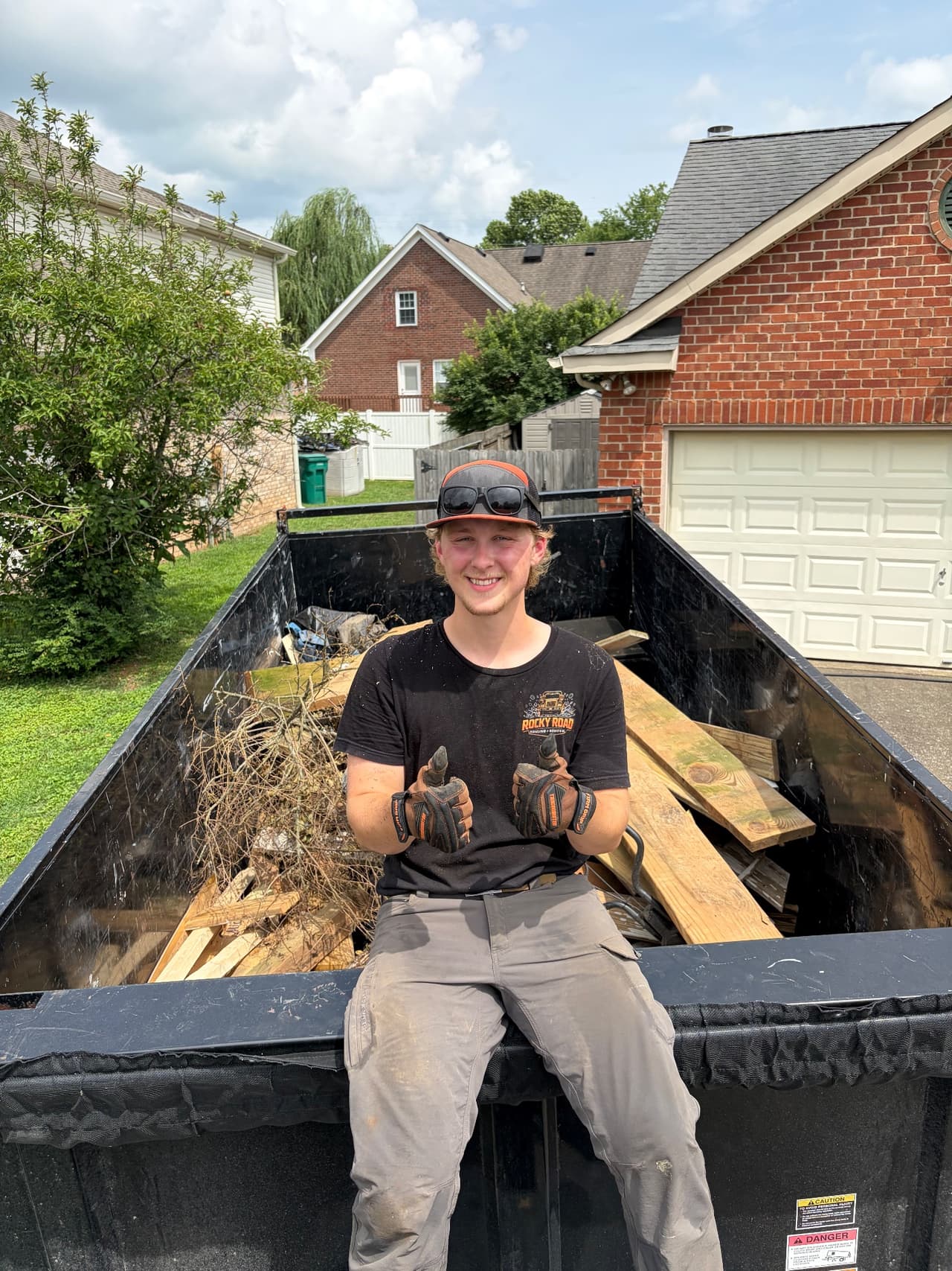 A Rocky Road crew member sitting on a loaded dumpster, giving a thumbs-up on a residential job site